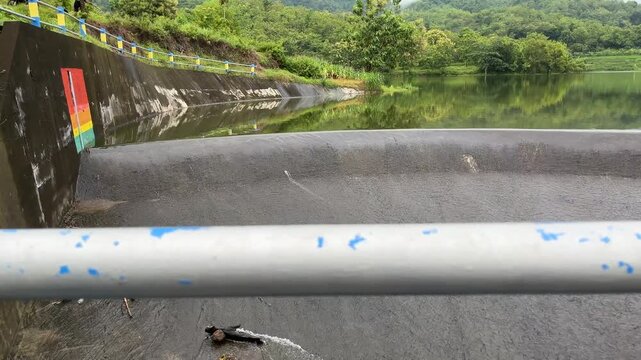 Foreground metal pipe framing a calm reservoir and dam landscape, blending industrial water infrastructure with natural scenery.