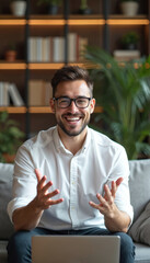 Fototapeta premium Smiling man with glasses in white shirt gestures talking during video call. He sits on couch near laptop in cozy home office with bookshelves and plants. Casual communication via computer.