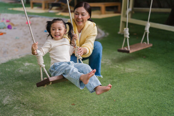 cheerful child girl with mother playing on wooden swing at playground