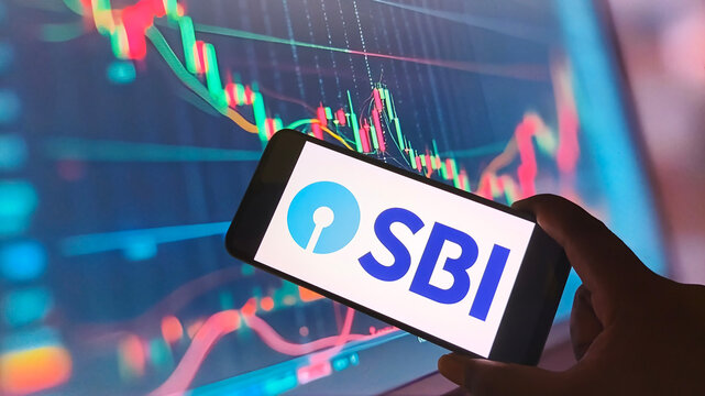 Hand holding smartphone with SBI logo on screen in front of stock market graph display