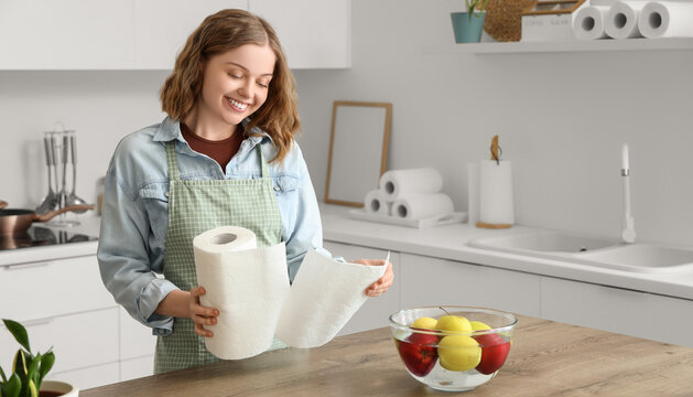 Young woman with roll of paper towels and apples near table in kitchen