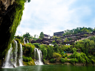 Scenic view of Furong Ancient Town in China, nestled on a green cliff with cascading waterfalls and traditional wooden houses, perfect destination for eco-tourism, culture, and historic architecture.