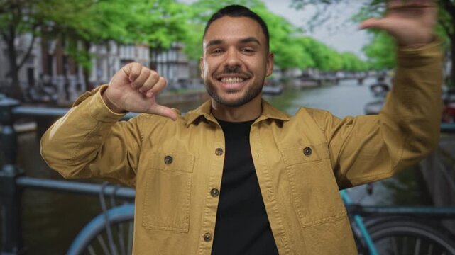 Man in yellow jacket smiling and framing with hands on busy street in city beside canal railing and bicycle wheel; joy.