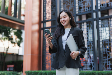 Young businesswoman smiling holding laptop using smartphone outdoors