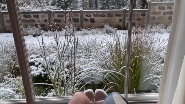 Pov from a rocking chair looking down at feet in slippers on a footstool and hands on a blanket, with slow snowfall in a windless winter garden outside the window, concept of gentle motion 