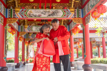 Fototapeta premium Happy senior Asian couple holding red shopping bags at Chinese temple during Lunar New Year, Elderly husband and wife laughing in cheongsam celebrating shopping festival. Retirement lifestyle concept