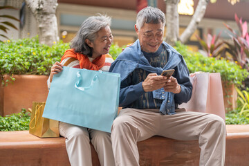 Happy senior Asian couple sitting on bench with shopping bags, Elderly husband using smartphone while wife leaning on shoulder smiling, Retirement lifestyle and digital technology concept outdoors
