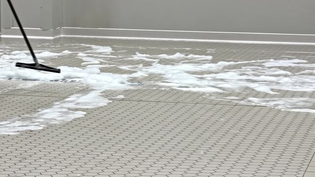 Worker pushes thick cleaning foam across tiled flooring with a floor squeegee, showing industrial sanitation, spill control, and professional wash-down maintenance in progress.