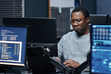 Black man, young adult, sitting at desk working on laptop surrounded by multiple computer monitors displaying code in modern office environment, focused on screen