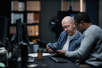 Middle aged Caucasian man and young adult Black man working together at desk using digital tablets and computer monitors, focusing on documents and collaborating in modern office