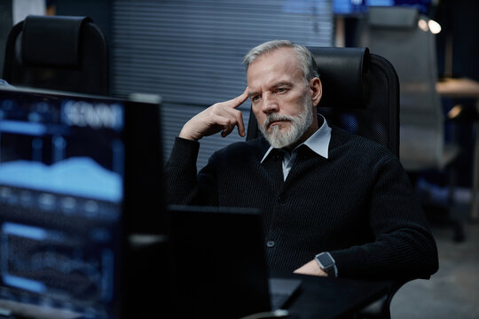 Middle aged Caucasian man sitting at desk thinking deeply, resting head on hand, looking at computer monitors in modern office environment, appearing focused and contemplative