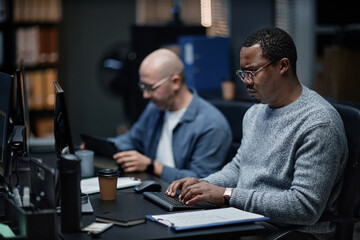 Middle aged Black man working on computer at desk beside middle aged Caucasian man using digital tablet in modern office setting, both focused on tasks, technology visible on desks