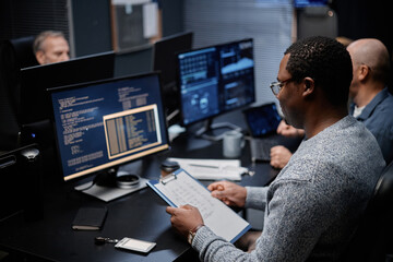 Black young adult man sitting at desk analyzing code on computer monitor while holding clipboard with notes, working alongside middle aged Caucasian men in modern office setting