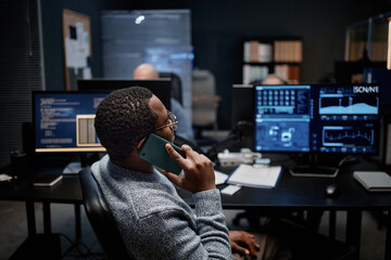 Black young adult man sitting at workstation talking on smartphone while monitoring data on multiple computer screens in modern office, another middle aged man working in background