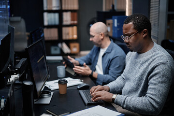 Middle aged Black man working on computer at desk in modern office, sitting next to middle aged Caucasian man using digital tablet, both focused on technology tasks in professional workspace
