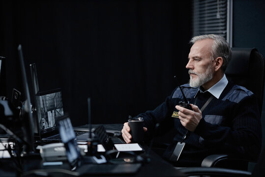 Middle aged Caucasian man sitting at desk, monitoring security cameras, holding walkie talkie and coffee cup, wearing security uniform focused on multiple computer screens in control room