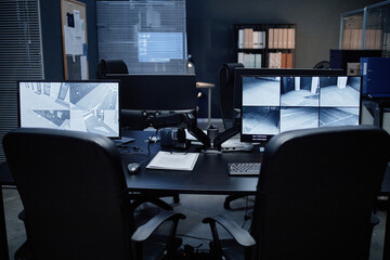 Naklejka premium Empty security office showing two computer monitors displaying multiple surveillance camera feeds, office chairs positioned in front of desk, various documents and equipment arranged on tabletop