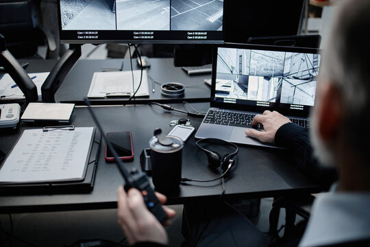 Middle aged Caucasian man monitoring security camera feeds on laptop and multiple screens while holding walkie talkie, working at surveillance control center with electronic devices
