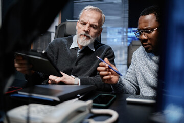 Middle aged Caucasian man and young adult Black man collaborating at desk, examining digital tablet and discussing information, both focused on technology in modern office setting