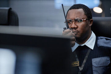 Black middle aged man wearing glasses operating walkie talkie sitting at security desk focusing on monitoring screens during surveillance shift in control room