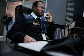 Black middle aged man wearing security uniform sitting at desk, talking on walkie talkie, holding identification badge, working in control room with computer monitors visible in background