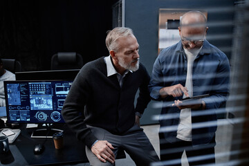 Middle aged Caucasian man sitting on desk leaning forward listening to bald man using digital tablet, both collaborating in modern office with computer monitors displaying data