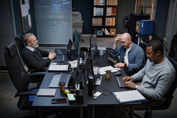 Three middle aged men, including Caucasian and Black individuals, working at desks with laptops and documents in modern office, focusing on tasks and collaborating on project