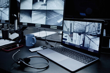 Multiple surveillance camera feeds displaying on computer monitors and laptop screen, electronic devices including smartphone and headset resting on dark desk in security control room
