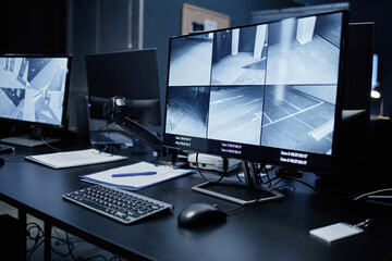 Fototapeta premium Empty security control room showing modern surveillance workstation with multiple monitors displaying live CCTV footage of building corridors and parking areas, keyboard and documents on desk