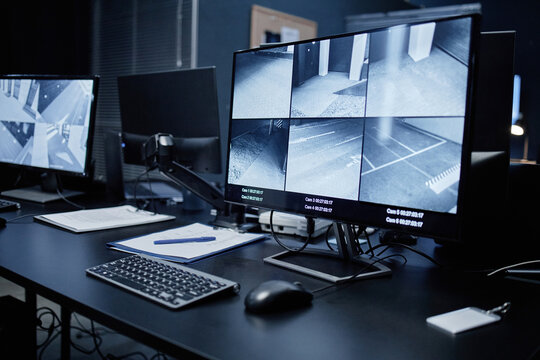 Empty security control room showing modern surveillance workstation with multiple monitors displaying live CCTV footage of building corridors and parking areas, keyboard and documents on desk