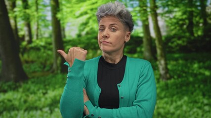 Woman with short gray hair wearing green cardigan and black top points thumb at forest backdrop indoors; skepticism doubt cynicism distrust.