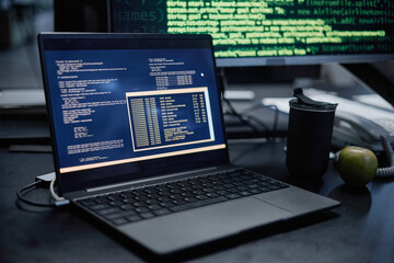 Laptop displaying programming code on screen sitting on desk with reusable cup and green apple nearby, computer monitor in background showing green text on black background