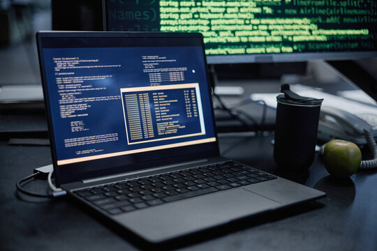 Laptop displaying programming code on screen sitting on desk with reusable cup and green apple nearby, computer monitor in background showing green text on black background - Powered by Adobe