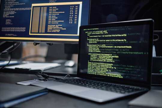 Closeup showing laptop with green code on screen positioned in front of desktop monitor displaying command line interface, illustrating software development or cybersecurity workspace