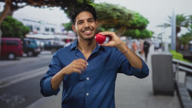 Young hispanic man wearing blue shirt holds small red heart and forms heart with hands on street; love joy warmth.