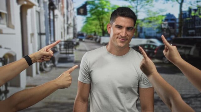 Man in white t shirt on street with multiple fingers pointing to him from four hands, neutral expression; judgment social pressure.