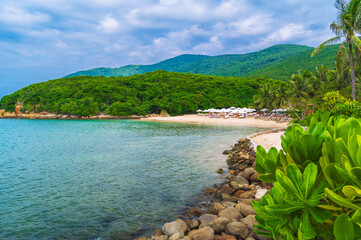 sandy beach with sun umbrellas and sun beds under palm trees in bay by sea on cloudy summer day at a resort on island