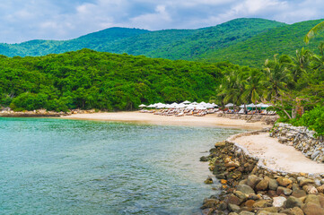 landscape with sandy beach with sun umbrellas and beds under palm trees in bay by sea on cloudy summer day at resort on paradise island