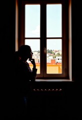 Contemplative Silhouette of a Woman Standing by a Window with Urban Scene in Background, Bathed in Subtle Indoor Light, Depicting Solitude and Introspection