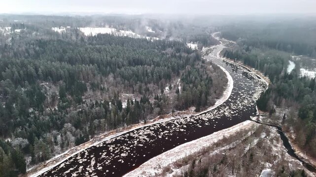 Aerial view of a winding winter river flowing through dense snowy forest, with drifting ice patches and misty cold air above the landscape.