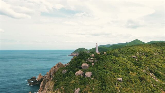 Cinematic drone shot of the historic white lighthouse at Mui Dai Lanh, perched on lush green mountains overlooking the blue East Sea under a bright sky.