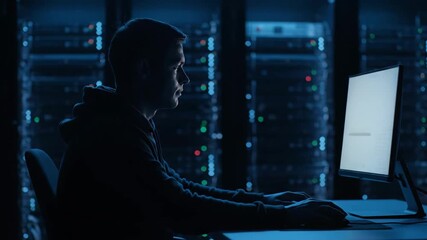 A young man works on a computer in a dark server room with glowing server racks. Represents cybersecurity and IT. - Powered by Adobe