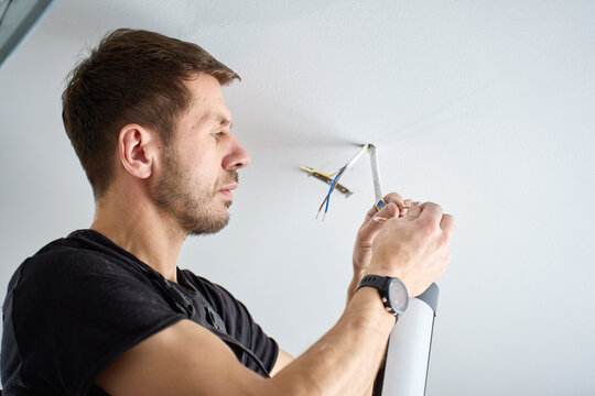 Man in workwear connecting electrical wires while installing ceiling light fixture indoors. Concept of lighting installation, electrical work and home interior repair
