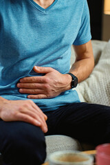 Man sitting on sofa in living room clutching stomach and suffering from abdominal pain or cramps. Person suffering from health issues, digestion problems or food poisoning at home