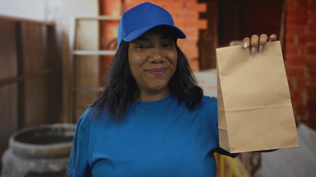 Woman wearing blue cap and blue shirt points finger to a brown paper bag at an indoor construction site; confidence trust.