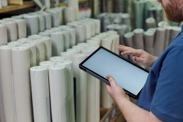Caucasian man using digital tablet while standing in warehouse, checking inventory of large rolls of material, focusing on screen and managing stock control process