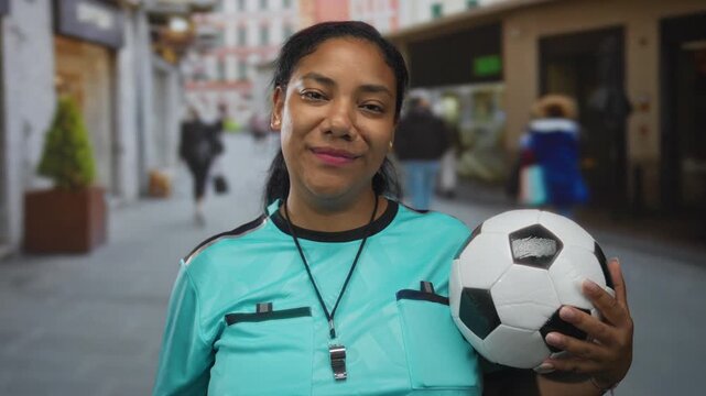 Woman referee in turquoise uniform holds soccer ball under arm and points fist forward on urban street; authority fairness impartiality control.