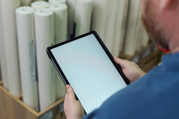 Middle aged Caucasian man holding digital tablet with blank screen standing in front of rolls of paper in warehouse, using device for inventory management or quality control