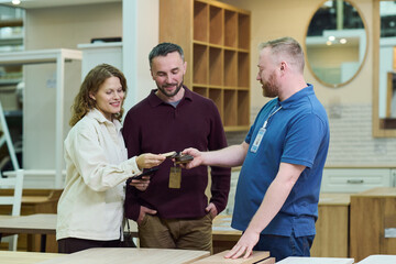 Fototapeta premium Caucasian young adult woman and Caucasian young adult man selecting wood samples with assistance from Caucasian young adult male store employee in furniture showroom
