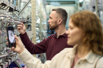 Caucasian man examining faucet fixtures on store shelf while Caucasian young adult woman taking photo with smartphone in foreground, both shopping in hardware store aisle